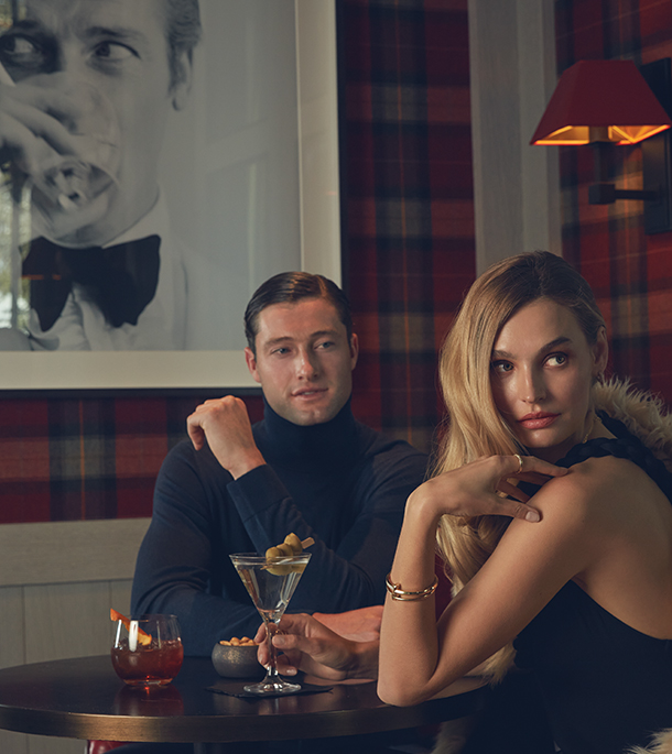 Man wearing dark turtleneck sweater and woman wearing a black dress, gold bangle and ring sitting at a table with cocktails