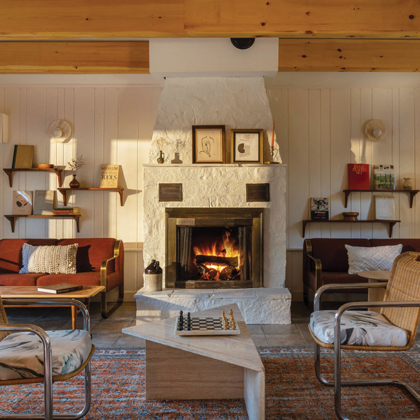 A cozy lounge area at a hotel with natural light coming in. The room has wooden beams on the ceiling, a fireplace, couches, chairs, bookshelves, a chess board on a coffee table.