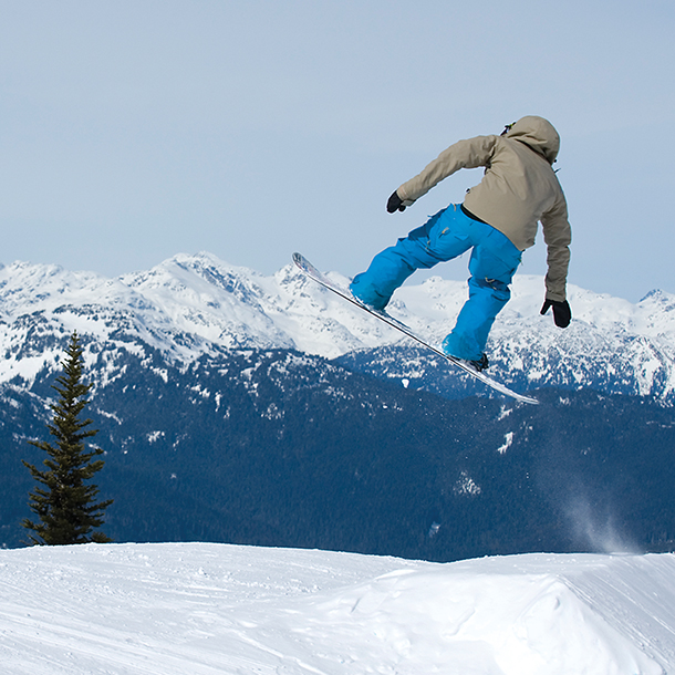 A snowboarder wearing a brown snow jacket and blue pants is doing a jump over a snow-covered slope. There are tress and snow-covered mountains in the background.