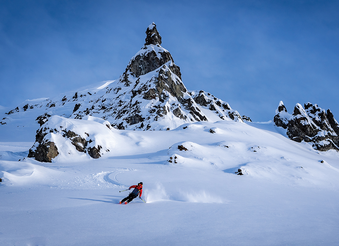 A heli-skier wearing red and black ski clothes skiing downhill on a run from a snowy mountain peak