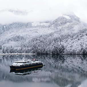 A catamaran boat floating on a frozen lake, surrounded by snow-covered trees and mountains.