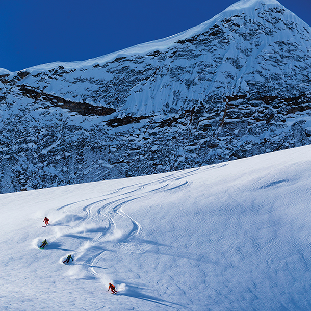 A group of people heli-skiing down a snow-covered slope on a sunny winter day. The skiers are all wearing colorful ski jackets and pants. The snow is sparkling in the sunlight, and the mountains in the background are majestic.