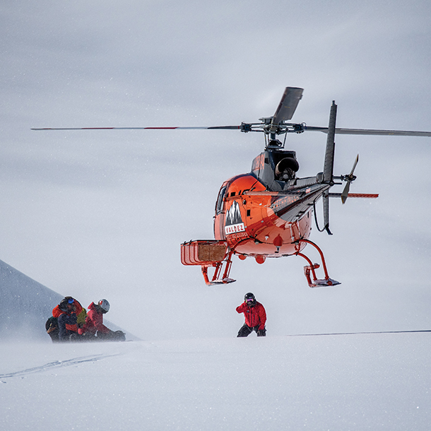 An orange helicopter bringing down heli-skiers to a snowy summit. Heli-ski guides wait in the snow.