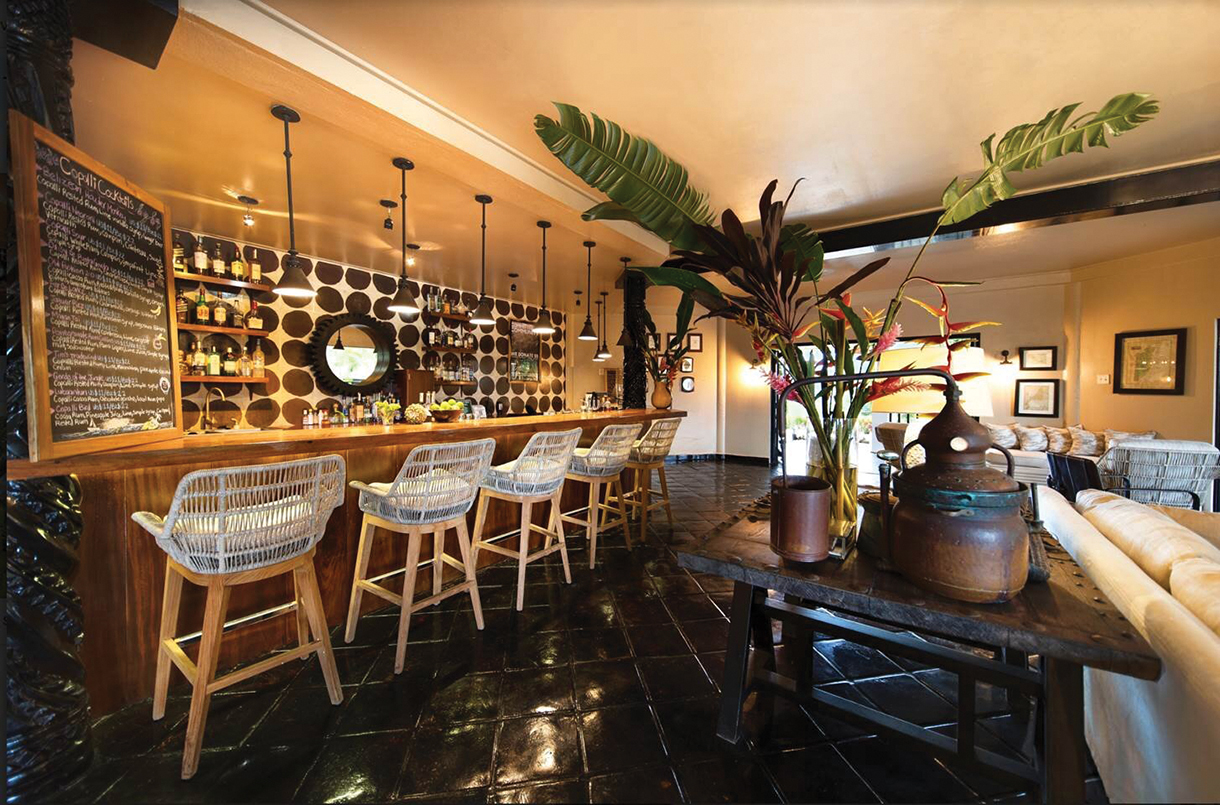 A bar interior with furniture including stools, console table and couches. Behind the bar, there is a large wooden shelf displaying various bottles of alcohol. 