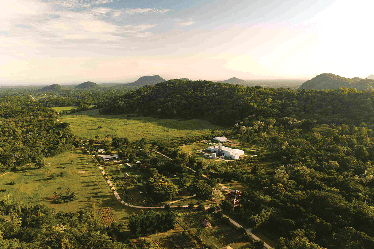 Aerial view of a lush green valley with a farm, road and white distillery building in the center. There are mountains in the background.