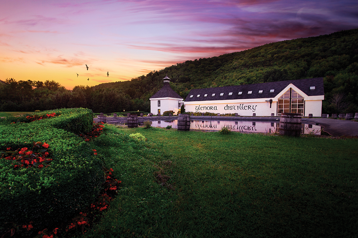 A large white building with a dark roof. The building has several stories and many windows. There is a green hillside behind the distillery, and trees in the distance.