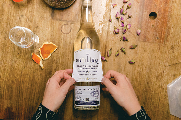 A person putting on a label onto a glass bottle of gin. The bottle is on top of a wooden table with an empty glass next to it.