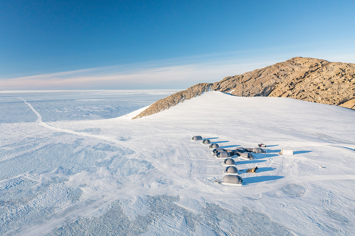 Gray, space-inspired, bubble accommodation pods at a remote base camp in Antarctica’s pristine white interior mountain range. Brown pitted rock formations protrude from Henriksen Nunataks’ glaciers.