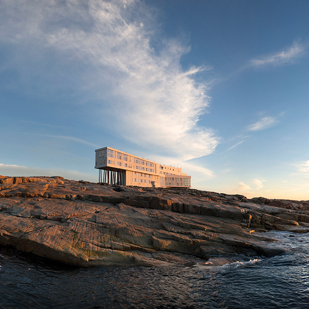 A large, contemporary building perched on stilts on one side, sits on top of a rocky hill that overlooks the ocean. The building is primarily made of wood and glass and has floor-to-ceiling windows.