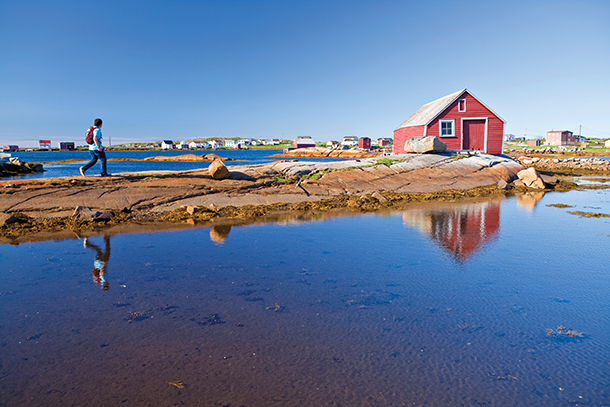 A man wearing a backpack walking on a path alongside a calm body of water. There is a wooden red house with white trim in the distance against the backdrop of a clear blue sky.