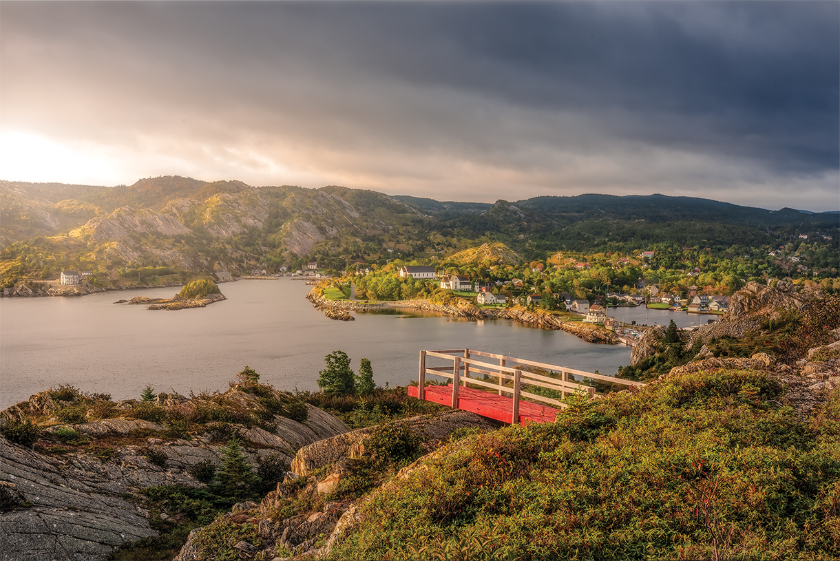 A red wooden deck juts out from a rocky hilltop, overlooking stunning views of a vast, sparkling lake surrounded by lush greenery.