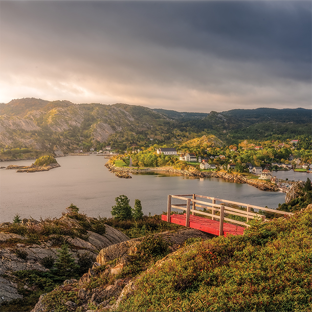 A red wooden deck juts out from a rocky hilltop, overlooking stunning views of a vast, sparkling lake surrounded by lush greenery.