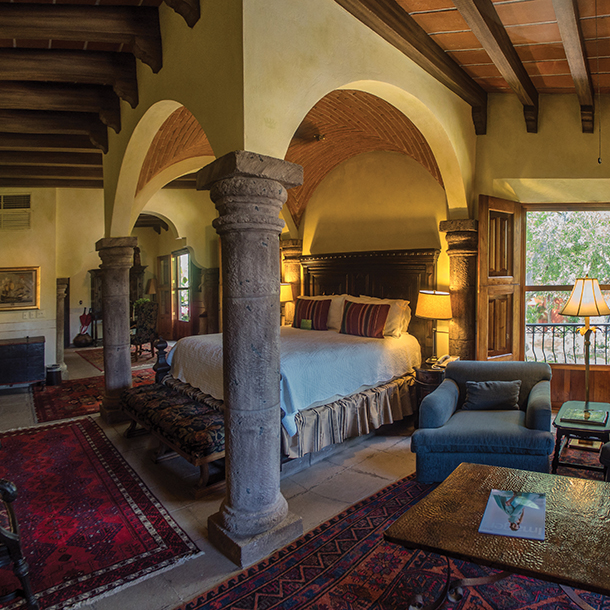 A guest suite with a large poster bed, table, chairs, balcony and fireplace in the background. The ceiling has wooden beams and the room is painted a mustard yellow. 