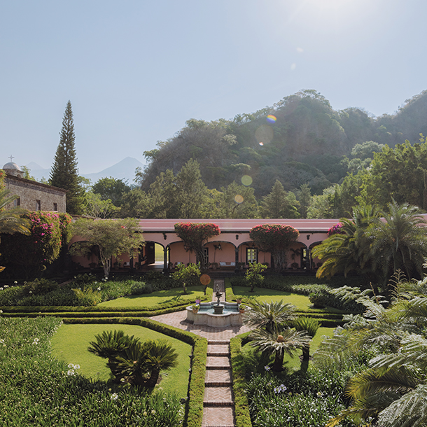 A lush green garden with a fountain as its focal point. The manicured garden is in front of a hacienda-style building with a paved walkway in the foreground.