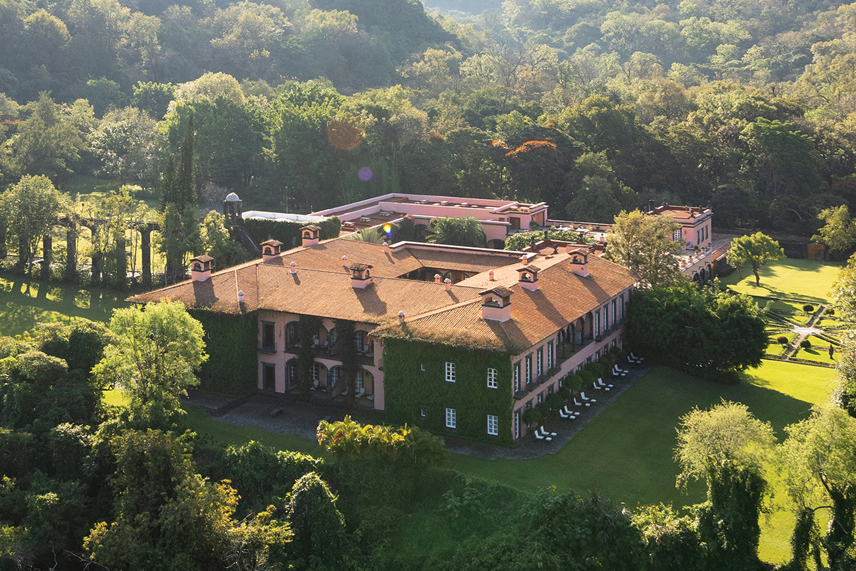 An aerial view of a large hacienda surrounded by lush, green highlands. The large estate has a red tile roof, many windows, and pink-colored stucco walls with some outer walls covered in greenery.