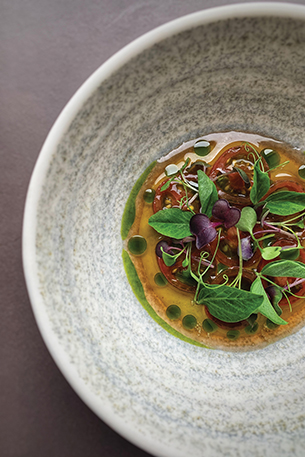 Bowl of tomatoes topped with a mixture of colorful edible leaves placed in a herb oil dressing.