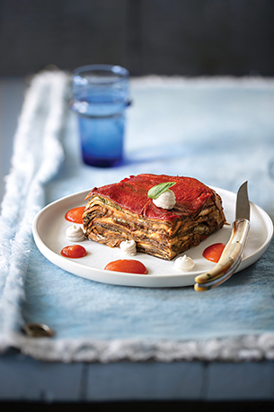 White plate with vegetable parmigiana on it. There is a knife placed on the plate and a blue drinking glass in the background on light blue table linens.