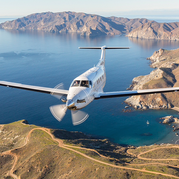 A small, white, airplane flying over a Pacific Ocean. In the distance, there is a long, low green mountain range.