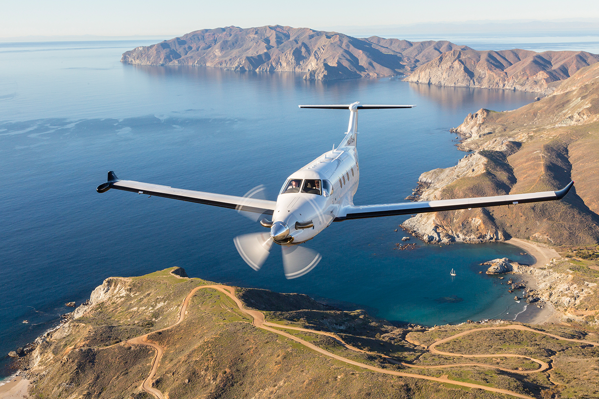 A small, white, airplane flying over a Pacific Ocean. In the distance, there is a long, low green mountain range.