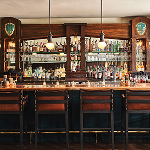 Roadside bar interior with a long wooden counter and several stools. There are many different sized bottles of alcohol lined up on shelves behind the counter.