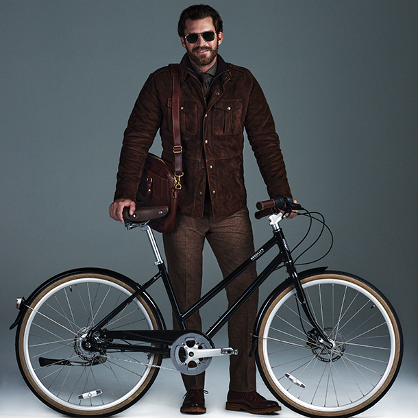 Man wearing black aviator sunglasses and carrying brown leather briefcase on his shoulder standing next to black Willow 7i Disc bicycle.