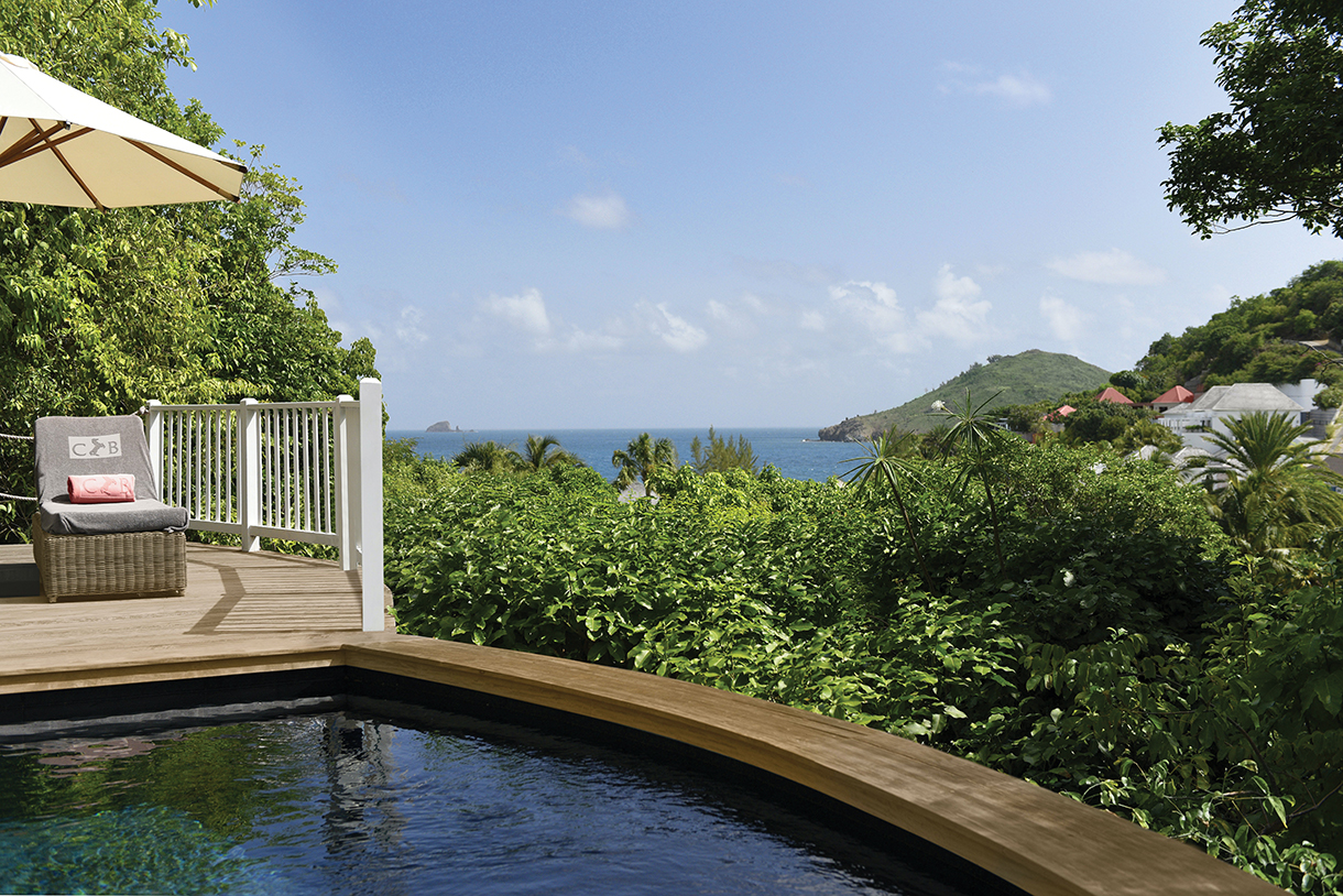 A swimming pool with a chair and umbrella on a deck overlooking the ocean and dense greenery