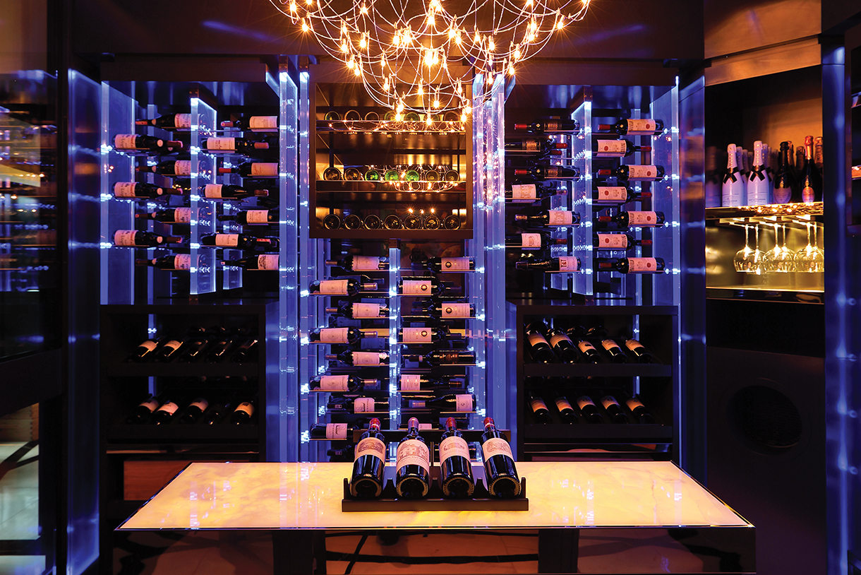 A dimly lit residential wine cellar with acrylic displays illuminated by controlled colored lights. The wine cellar has a shining chandelier, backlit onyx table and black lacquer frames which accentuate the glass-walled, cube-like architecture.