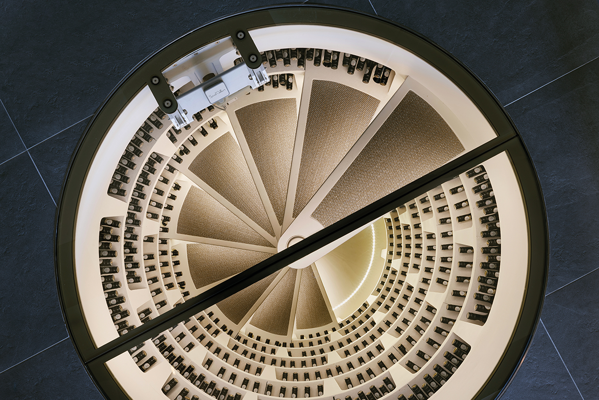 A below-ground wine cellar with wooden shelves stocked with bottles of wine. There is a spiral staircase with wrought iron railings that leads down into the wine cellar. Caption: Below-ground Spiral Cellar in London