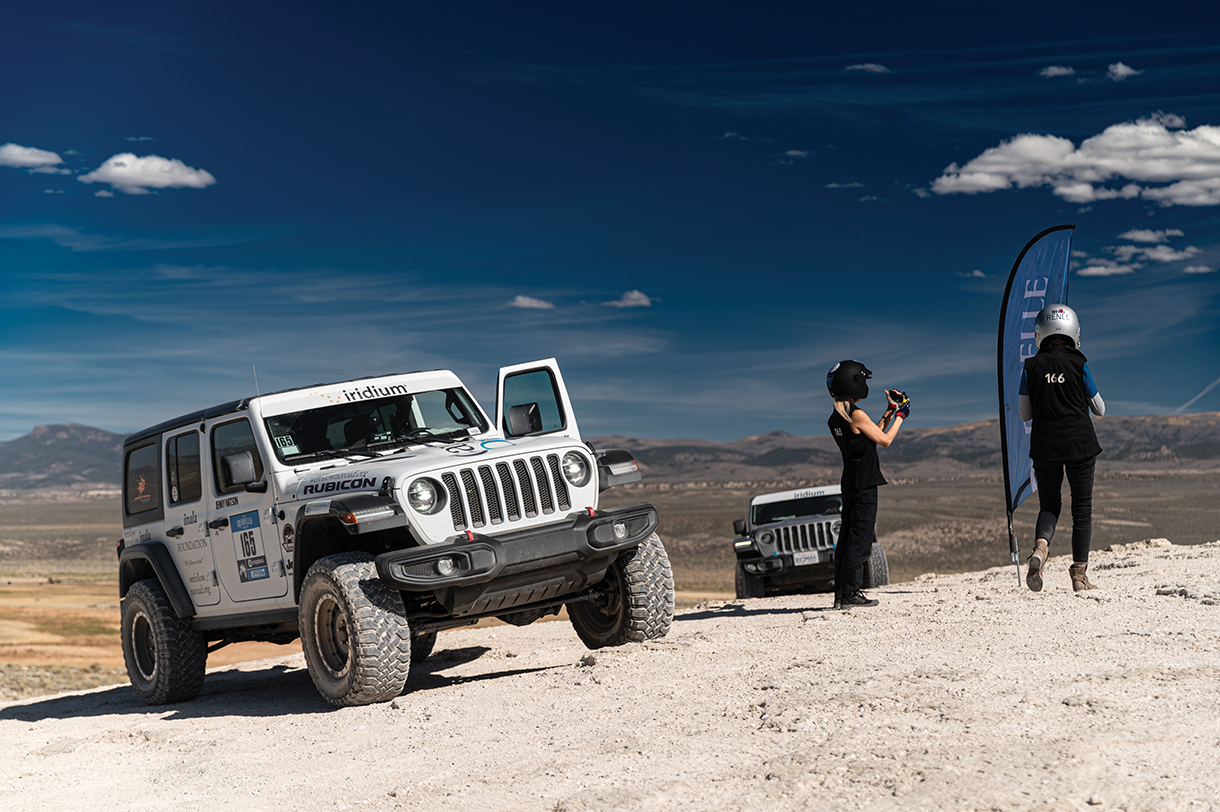 Two women wearing racing helmets stand outside parked electric vehicles to stop at off-road rally check point 