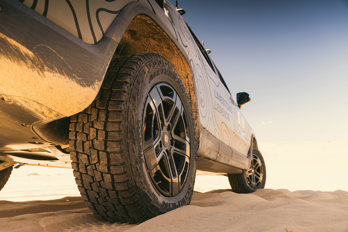 Closeup of the rear wheel, tire and side of gray Rivian R1T rally truck parked on sand