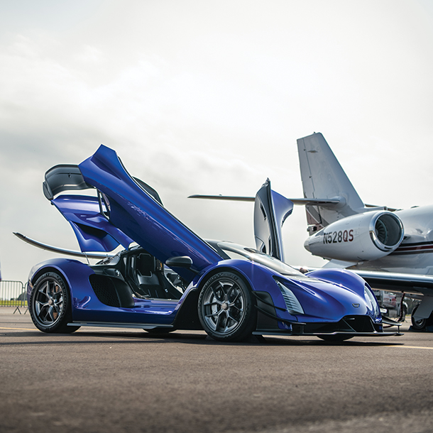 Front side view of blue CZinger with gullwing doors open and an aircraft parked next to it 