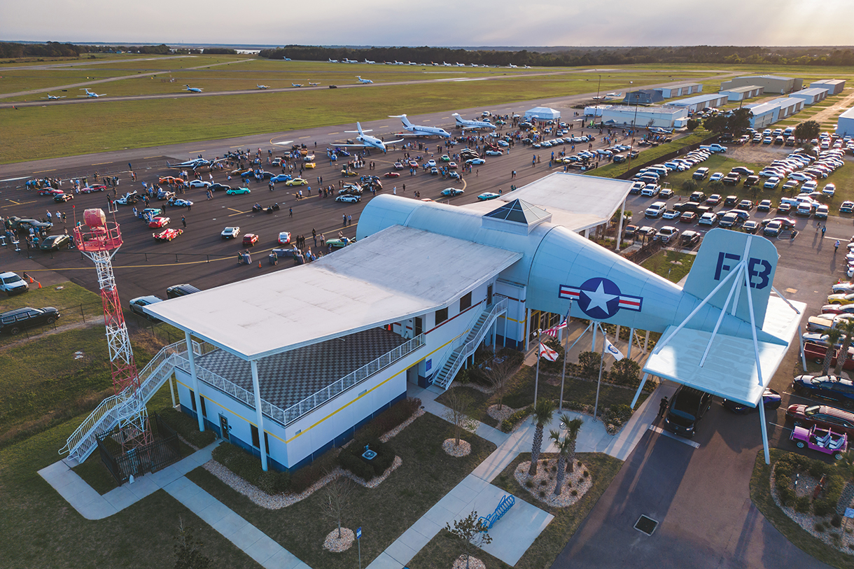 Top view of a car show at a historical airport that displays a vintage aircraft 