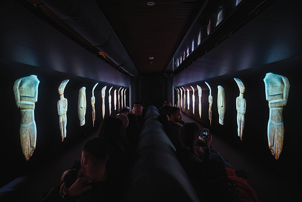 A museum exhibit featured in a dark, narrow room with a long, narrow bench running down the center. On either side of the bench are projections of ancient statues, some of which have human-like figures. The statues are illuminated in a soft, ethereal light. There are several people sitting on the bench.