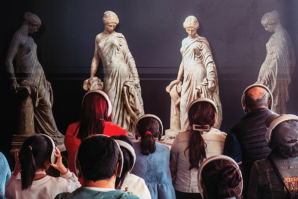 A group of people of different ages wearing headphones sitting down in front of four statues listening to a curator explaining an exhibition that highlights female figures in Mediterranean civilizations.