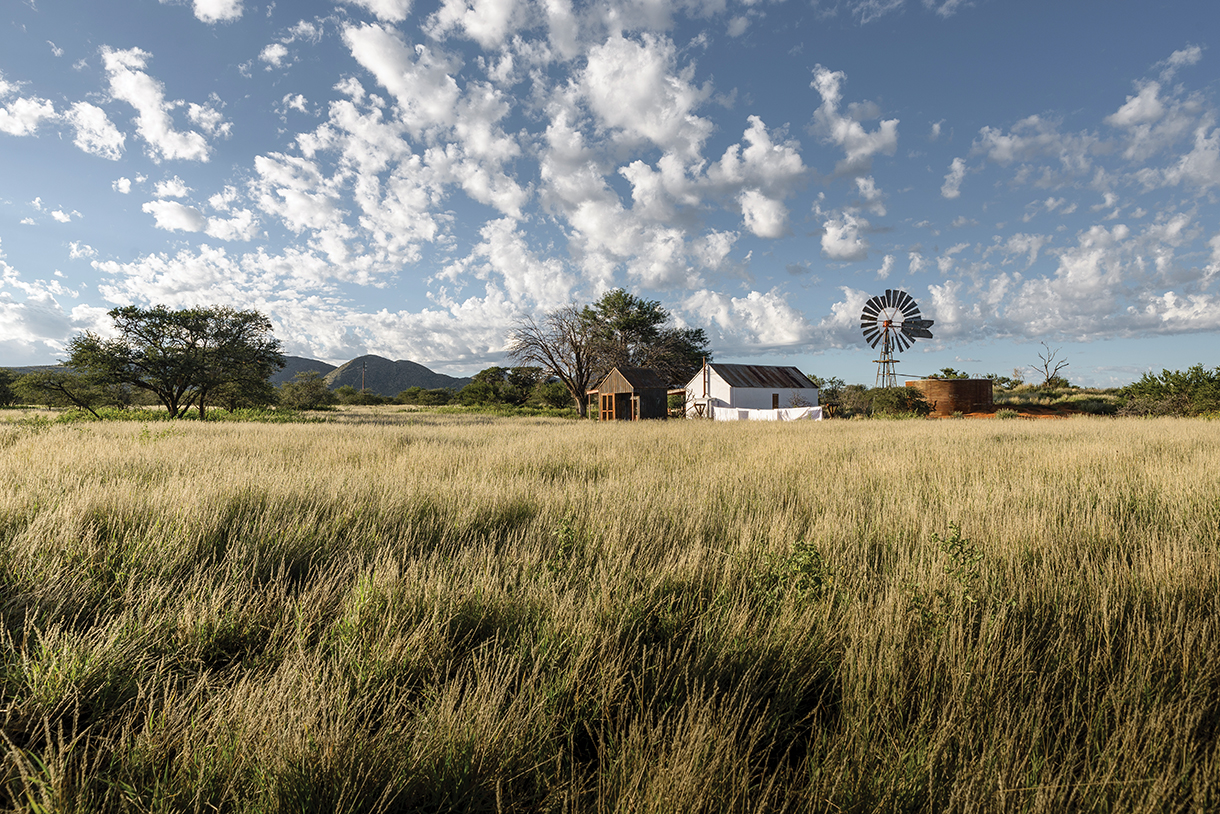 A rural landscape with a white farmhouse, a windmill, and a large water tank. The farmhouse is surrounded by tall grass and trees, with a blue sky and white clouds above.