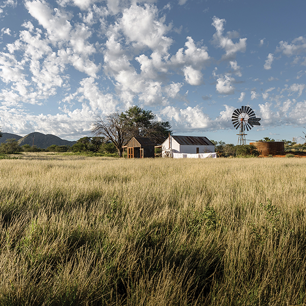 A rural landscape with a white farmhouse, a windmill, and a large water tank. The farmhouse is surrounded by tall grass and trees, with a blue sky and white clouds above.