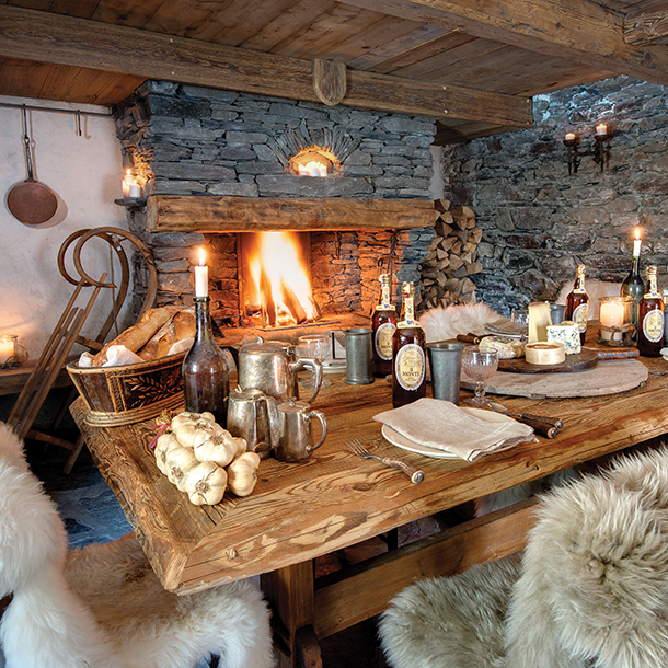 A cozy chalet dining room with a stone fireplace and a long wooden table set for a meal. Candles and lanterns illuminate the room, and there are fur rugs and a sled in the background.