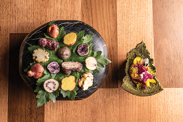  A plate of colorful native potatoes arranged on top of a large leaf on a black slate platter, accompanied by a small organic shaped green vessel with dessert and edible flowers.