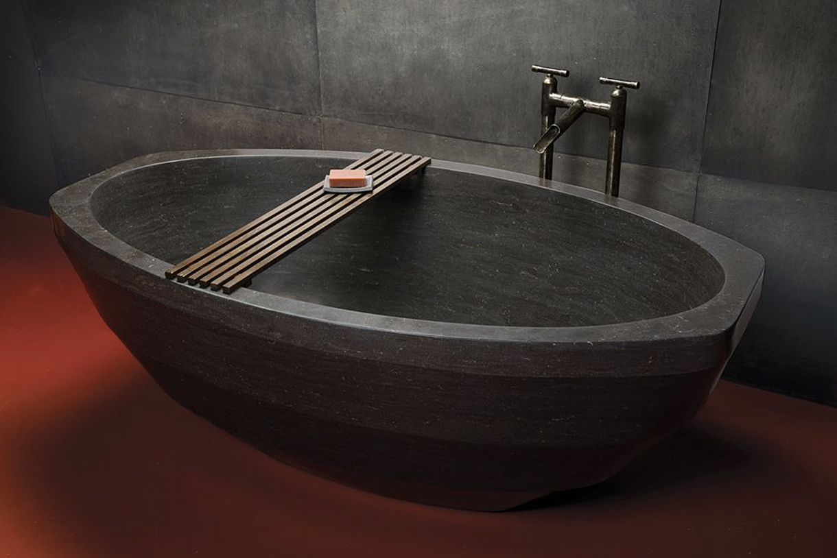 A large, oval bathtub made of antique gray limestone. The bathtub has a textured surface and a wooden shelf above it. A chrome faucet is mounted on the wall above the tub.