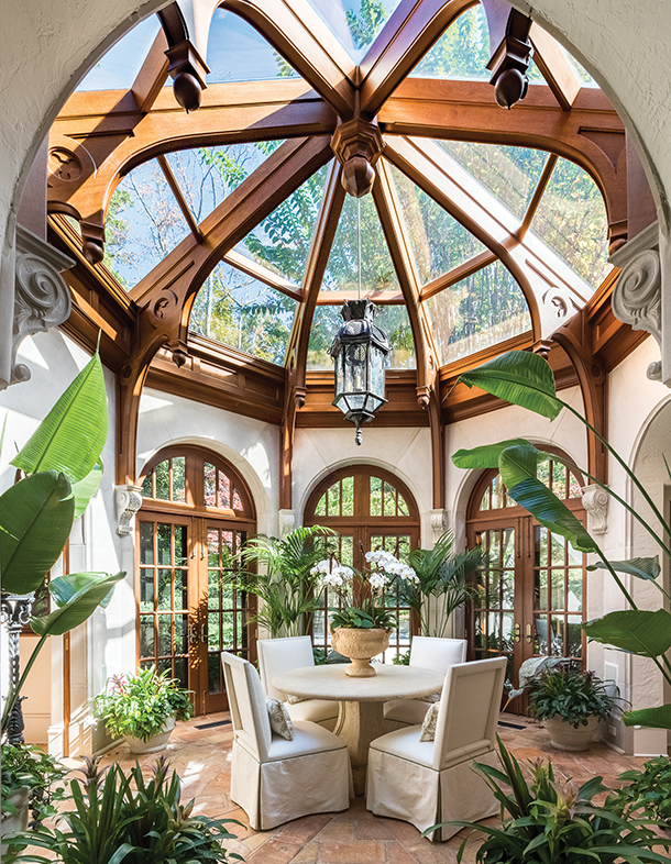 A sunroom with a domed glass ceiling supported by wooden beams. There is a round table with chairs in the center,surrounded by large potted plants. French doors lead to the outside.