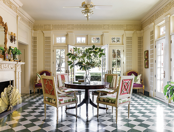 A spacious, sunlit room with a black and white checkered floor. There is a round table surrounded by four chairs, and a fireplace with a mantelpiece. French doors lead to a patio with a view of a garden.