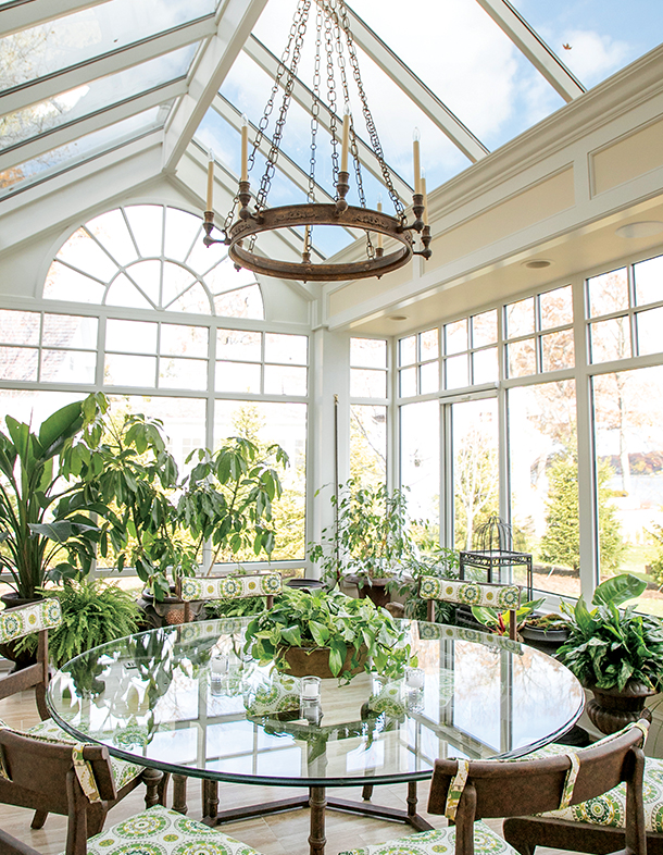 A sunroom with a glass ceiling and large windows, filled with various plants. A round glass table with chairs is in the center, and a chandelier hangs from the ceiling.