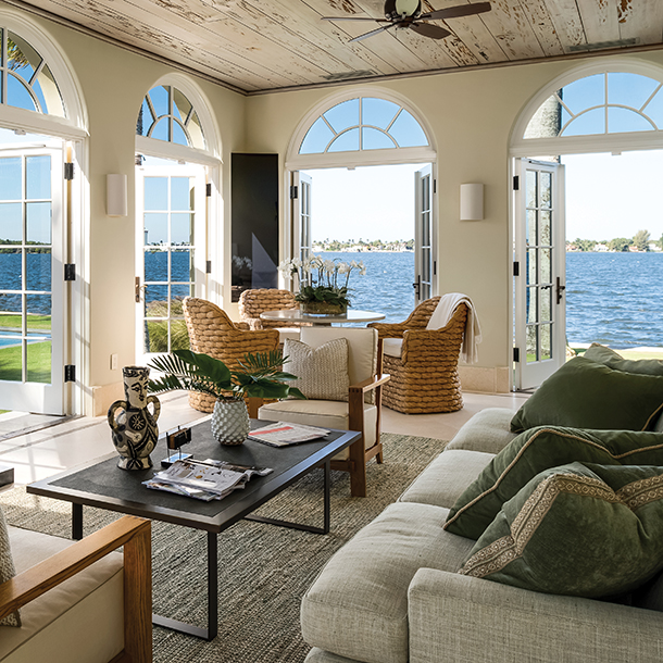 A bright and airy living room with arched windows overlooking a waterfront view. There is a sofa, coffee table, and armchairs arranged in a seating area. The room has a white ceiling with exposed wood beams and large glass doors leading to the outdoors.