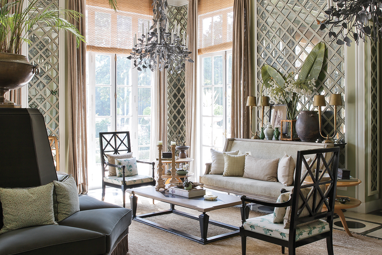 A spacious living room with large windows, a chandelier, and patterned wallpaper. There are several chairs, a sofa, and a coffee table in the room, and there are plants and vases as well.