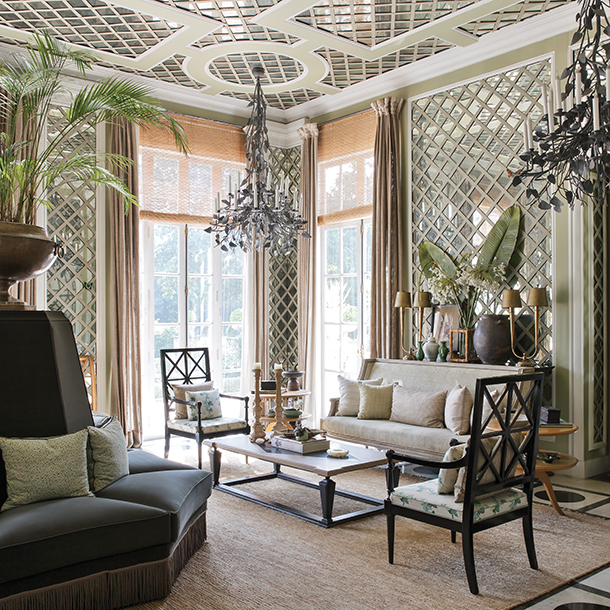 A spacious living room with large windows, a chandelier, and patterned wallpaper. There are several chairs, a sofa, and a coffee table in the room, and there are plants and vases as well.