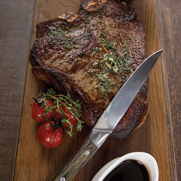  A perfectly cooked T-bone steak served on a wooden board with a side of roasted cherry tomatoes and thyme. A steak knife and a sauce boat with gravy are also on the board.