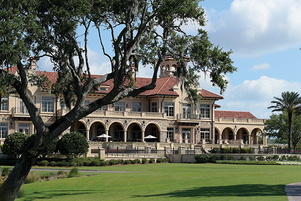 A grand clubhouse with a terracotta roof and arched windows, surrounded by lush green lawns and palm trees. A large oak tree stands in the foreground.