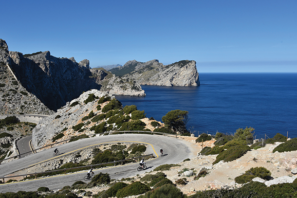 Overhead view of three cyclists riding on a narrow, winding paved road through rocky mountains next to a large body of water.