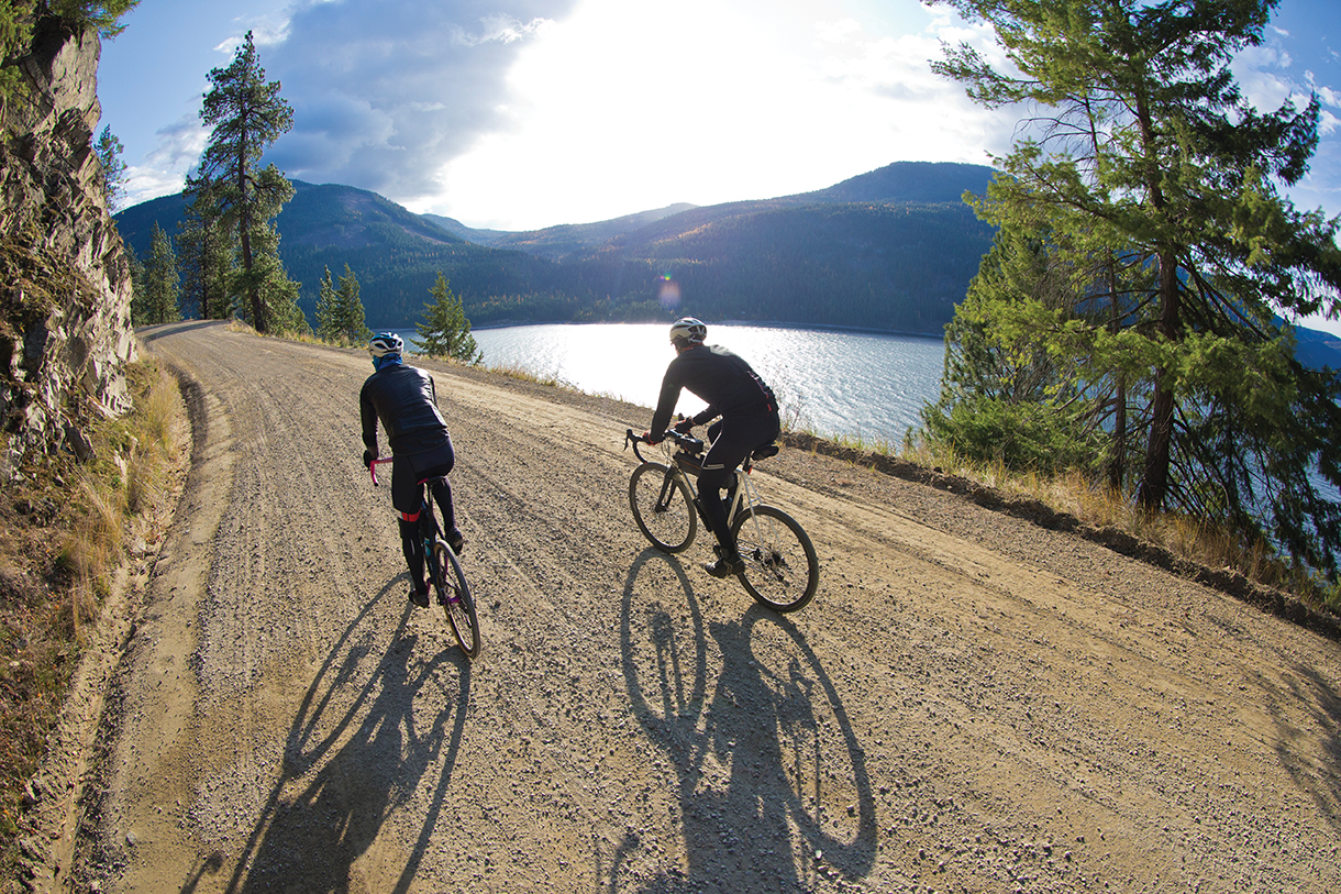 Two people riding bicycles on a gravel covered dirt road next to a rocky mountain and a body of water. There are grass covered mountains in the distance.