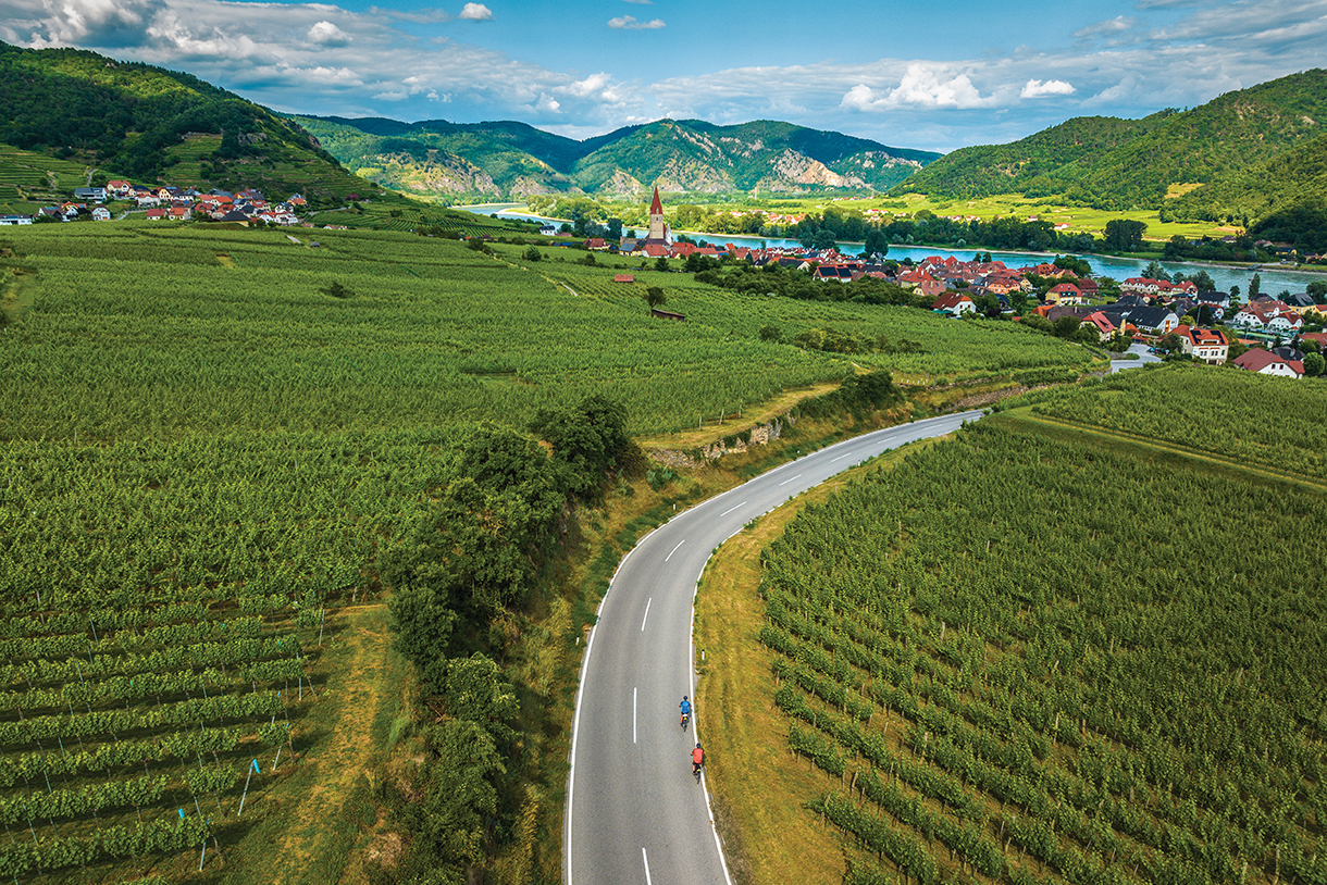 Aerial view of two people riding bicycles on a winding paved road through vineyards. There are houses, a river, and mountains in the background.